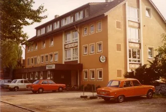 Ein altes Bild eines mehrstöckigen, gelb-orangefarbenen Gebäudes mit Fenstern und einer Markise. Vor dem Gebäude stehen mehrere Autos. Über dem Eingang ist ein Schild mit dem Wort "Wohnen" angebracht. Die Umgebung ist grün, mit Bäumen im Hintergrund.