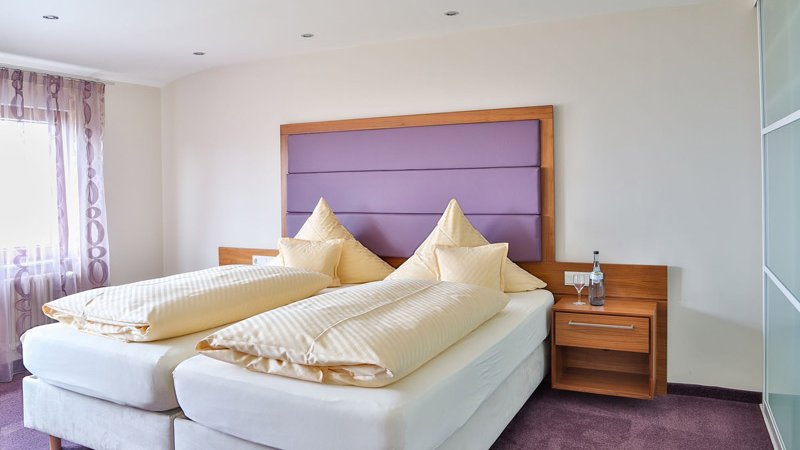 Ein Bett in der Theresia-Suite. Das Farbschema des Zimmers ist lila-braun. Rechts im Bild ist ein großer Glasschrank. 