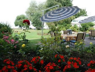Blick auf die blühende Gartenterrasse des Hotel Schwedi mit Sitzplätzen unter Sonnenschirmen, umgeben von roten Blumen und Blick ins Grüne.