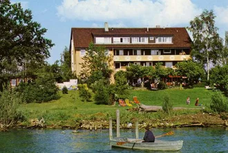 Ein ruhiges Seeufer mit dem zweigeschossigen Hotel im Hintergrund, umgeben von Bäumen und Grünanlagen. Im Vordergrund befindet sich ein kleines Boot mit einer Person, die rudert. Auf der Wiese sind einige Menschen zu sehen, die sich entspannen oder spielen.