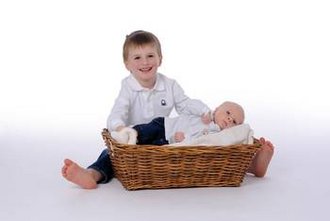 Das Kind Ben Göppinger sitzt auf dem Boden. Vor ihm liegt ein Körbchen mit dem Baby Tim Göppinger darin. Beide lächeln in die Kamera.