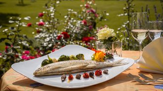 Elegant angerichteter Fisch mit Gemüse und Blütendekor auf Gartenterrasse des Hotel Schwedi – Feinschmeckergericht mit Blick auf Blumenwiese und Bodensee.