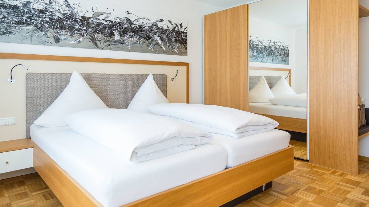 Ein großes Bett steht im Schlafzimmer der Franzl-Suite. Die Bettwäsche ist weiß, das Kopfteil und das abstrakte Bild sind in grauen Farben. Rechts neben dem Bett steht ein Holzschrank, in dessen Mitte ein großer Spiegel ist.