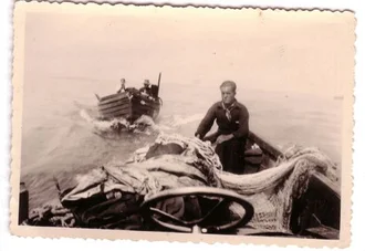 Ein Mann steht auf einem Boot und bedient das Steuer, während ein weiteres Boot im Hintergrund im Wasser fährt. Auf dem vorderen Boot sind Netze und Fracht sichtbar. Die Szene spielt sich auf einem Gewässer ab.