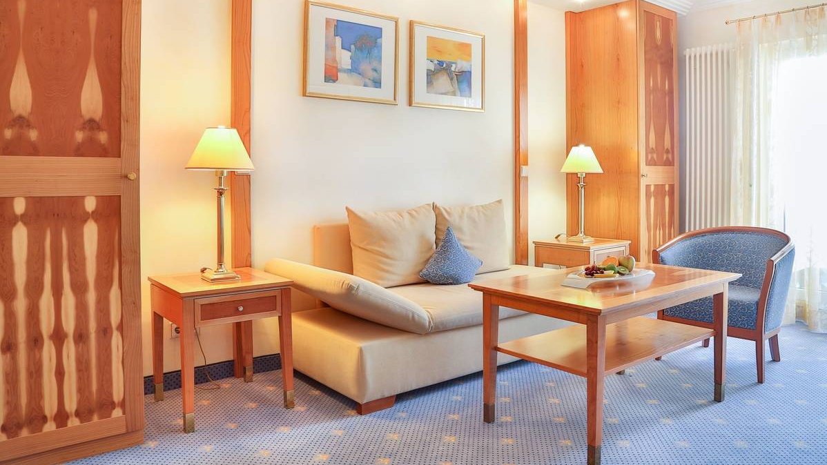 Wohnzimmer der Elisabeth-Suite. Darin befindet sich ein Zweisitzer-Sofa, ein Sessel, ein Couchtisch, zwei schmale hohe Schränke und zwei kleine Tische, auf denen gelbe Lampen stehen. Das Farbschema des Raumes ist braun-blau.