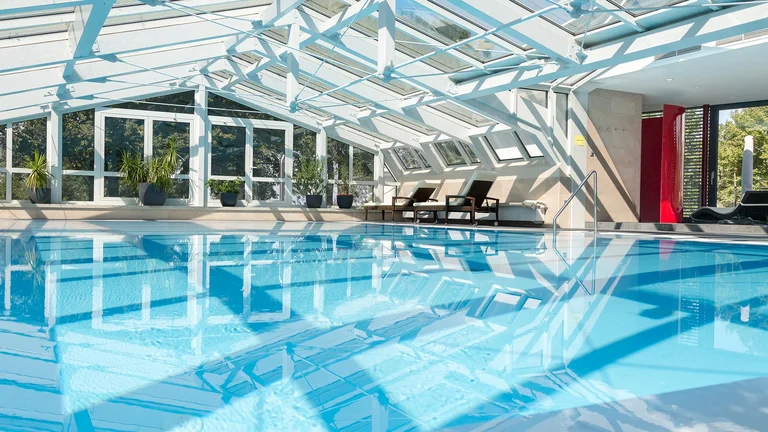 Ein großer, heller Innenpool mit klarem Wasser, umgeben von modernen Fenstern und Pflanzen. Die Architektur ist lichtdurchflutet, mit einem schrägen, transparenten Dach und gemütlichen Liegen am Rand des Pools.