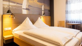 Ein großes Bett steht in einem Doppelzimmer. An der Wand hängt ein großes Bild auf dem Segelboote auf einem See zu sehen sind. Die kleinen Lampen neben dem Bett sind eingeschalten.