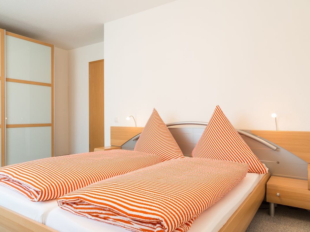 Schlafzimmer einer Ferienwohnung. Auf dem Bett liegt eine orange-gestreifte Bettwäsche. Links an der Wand steht ein Schrank mit Milchglas-Türen.