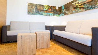 Zwei Sofas stehen in einer Ecke. Vor den Sofas stehen zwei quadratische, kleine Holztische. An der Wand hängt ein großes, längliches, abstraktes Bild.