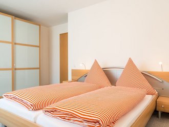Schlafzimmer einer Ferienwohnung. Auf dem Bett liegt eine orange-gestreifte Bettwäsche. Links an der Wand steht ein Schrank mit Milchglas-Türen.