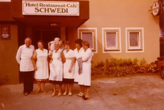 Eine Gruppe von sieben Personen steht vor dem Hotel-Restaurant-Café Schwedi. Alle tragen weiße Kleidung, einige in Koch- oder Kellneruniformen. Sie lächeln in die Kamera, während das Gebäude im Hintergrund sichtbar ist. Es sind Pflanzen und eine Einfahrt im Bild sichtbar.