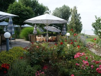 Blick auf die Gartenterrasse des Hotel Schwedi mit Sitzplätzen unter Sonnenschirmen, üppiger Blumenbepflanzung und Seepanorama – idyllischer Ort zum Verweilen im Freien.
