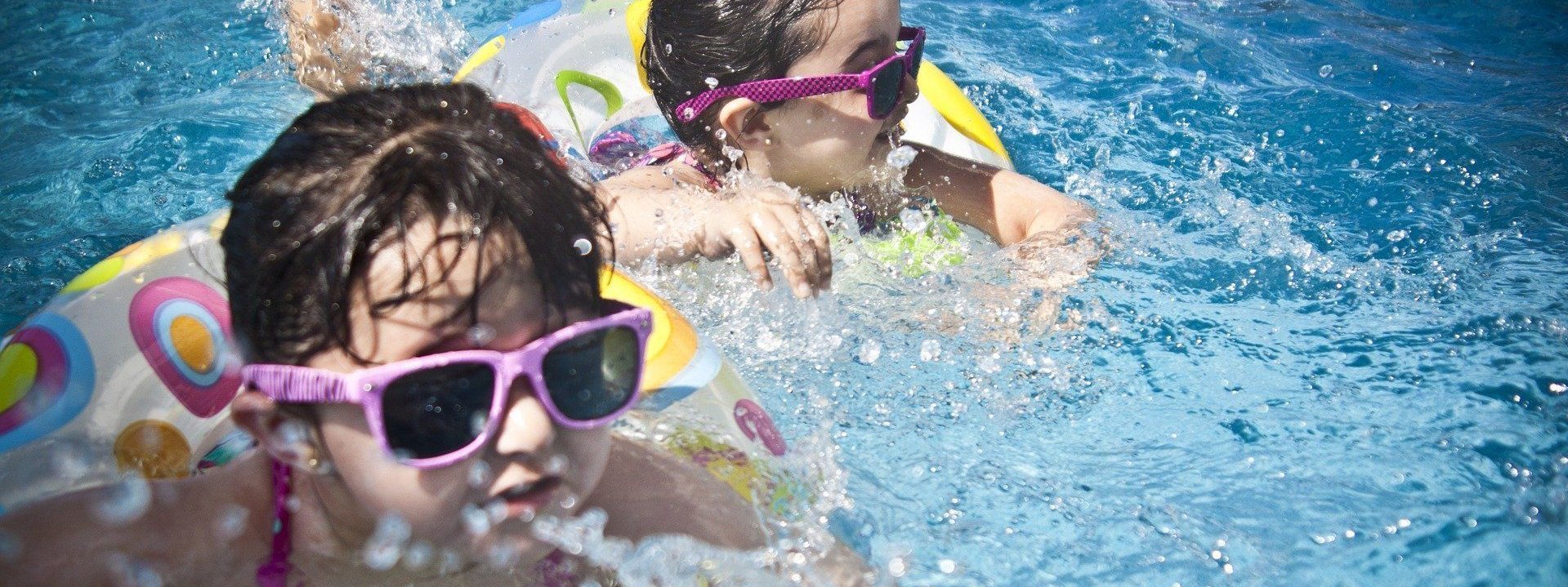 Zwei Kinder planschen mit Schwimmflügeln und Sonnenbrille im blauen Wasser.