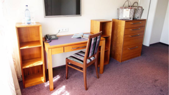 Auf dem Bild ist ein Schreibtisch mit Stuhl davor zu sehen. Links und rechts neben dem Tisch stehen zwei leere Regale, ganz rechts ist eine Kommode, auf dem zwei Körbe stehen.
