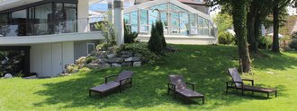 Das Hotel Schwedi mit großen Fenstern und einem Wintergarten steht auf einem grünen Rasen. Vor dem Haus befinden sich vier Liegen aus Rattan. Im Hintergrund wachsen Bäume, und es herrscht sonniges Wetter.