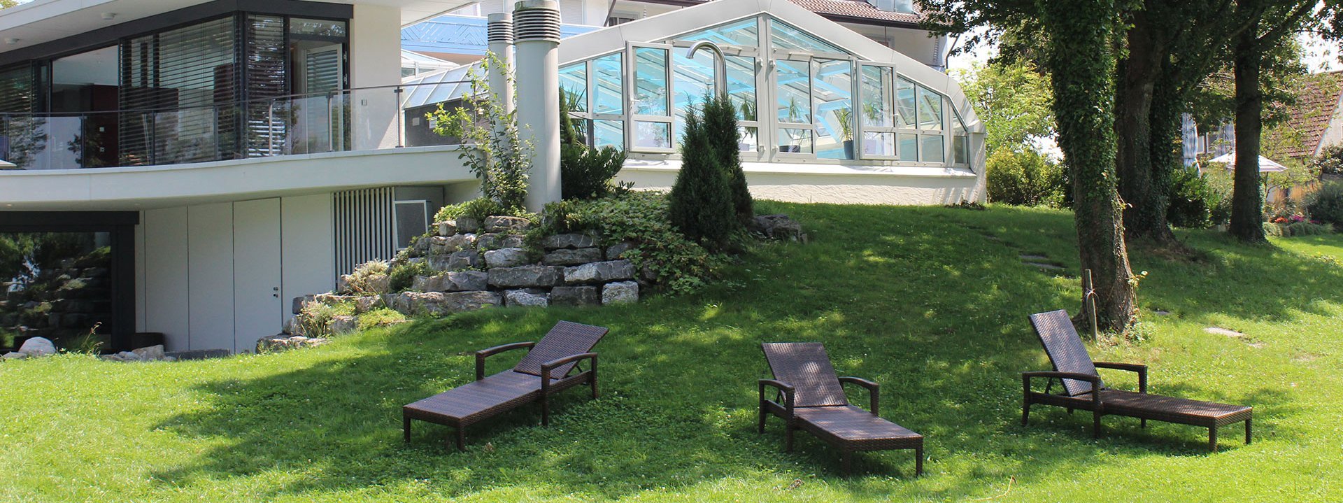 Im Garten des Hotel Schwedi stehen 3 Liegestühle. Teilweise befinden sie sich im Schatten der Bäume. Im Hintergrund ist der Pool zu erkennen.