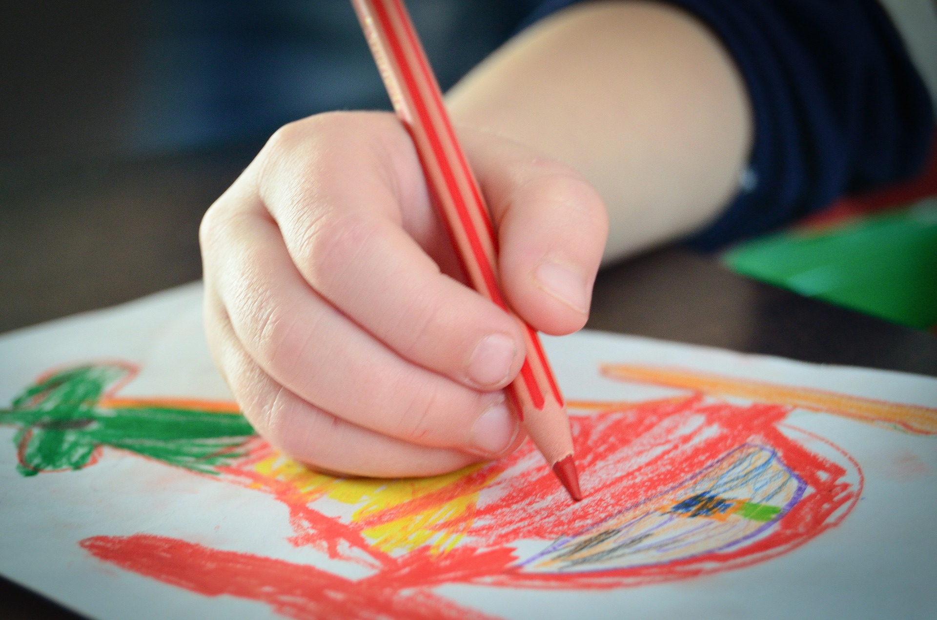 Eine Kinderhand hat einen roten Stift in der Hand und malt ein buntes Bild.